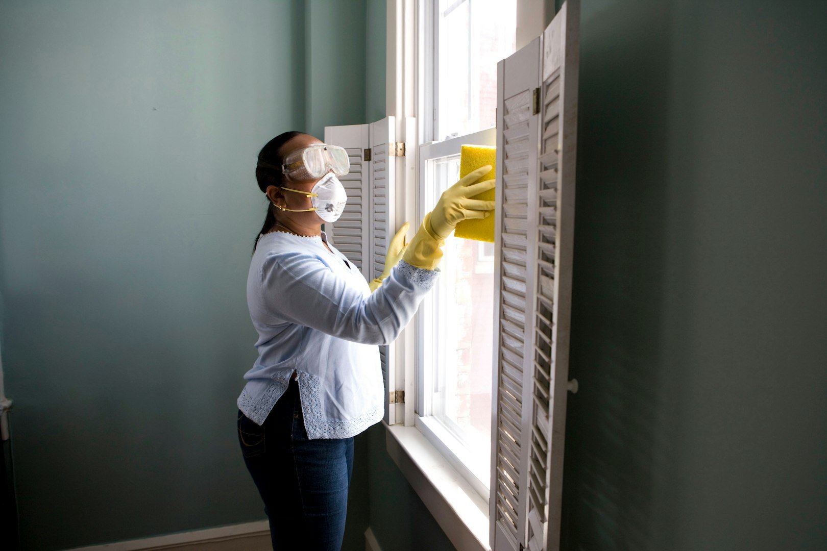 Three individuals in work uniforms and gloves are cleaning an indoor space with plants. One person is using a vacuum cleaner, while the others are engaged in cleaning tasks nearby. The setting features a modern interior with greenery and metal accents.